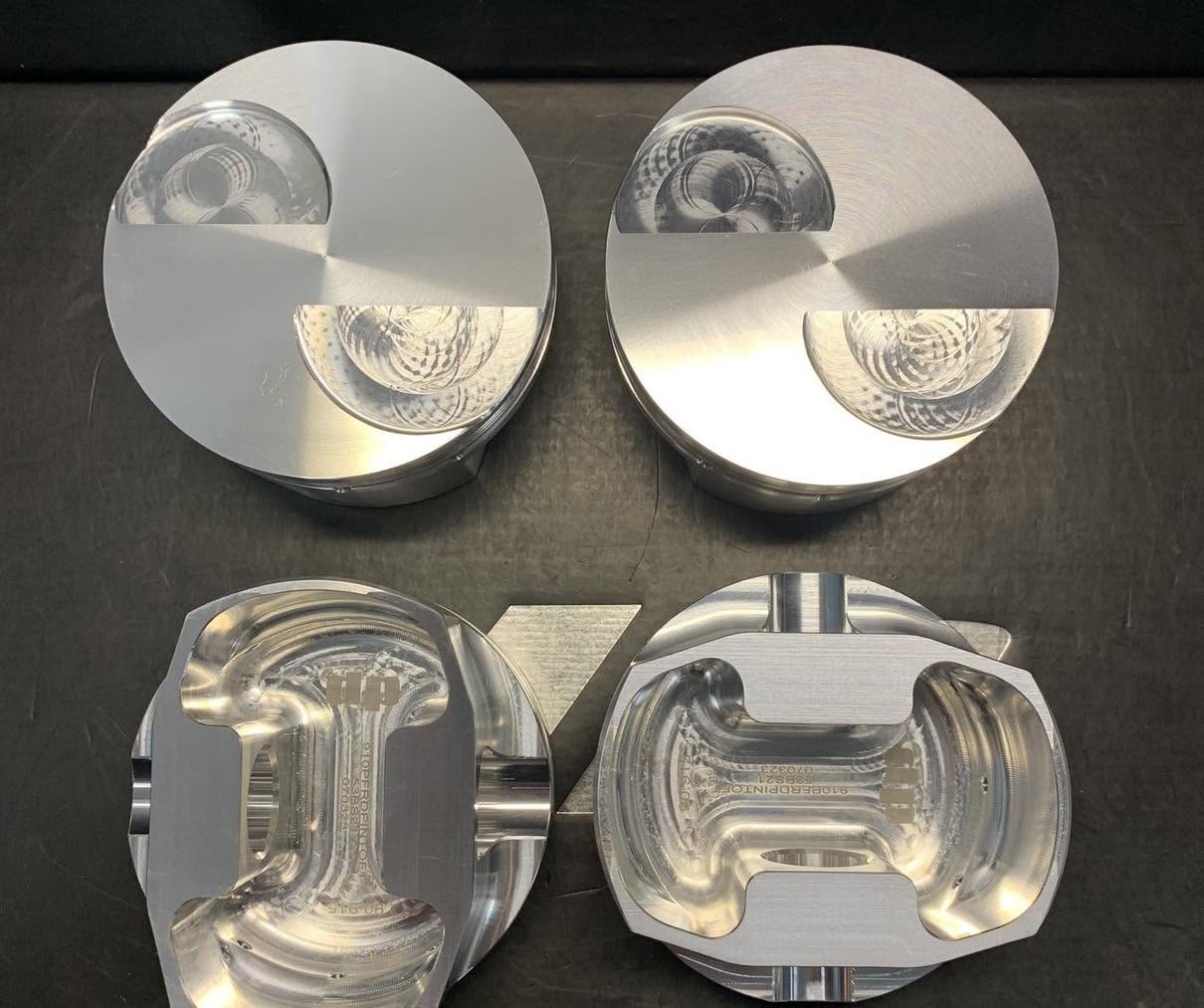 Top view of four aluminum pistons on a dark work surface: two complete pistons above, two cutaway pistons below showing inner chambers.
